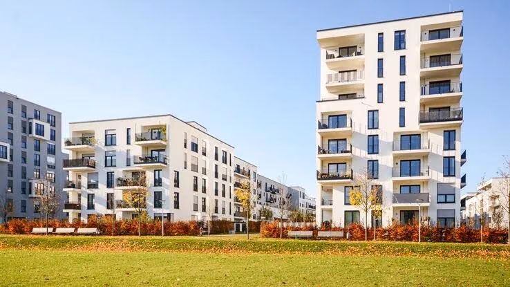 Das Foto zeigt mehrere Wohnblöcke mit modernen Wohnungen. Im Vordergrund ist ein kleiner Park mit Bänken, Bäumen und einer Wiese zu sehen.
