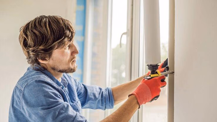 Ein Mann modernisiert seine Fenster. Er trägt Handschuhe und hält ein Werkzeug, mit dem er etwas zwischen Fenster und Wand gibt.