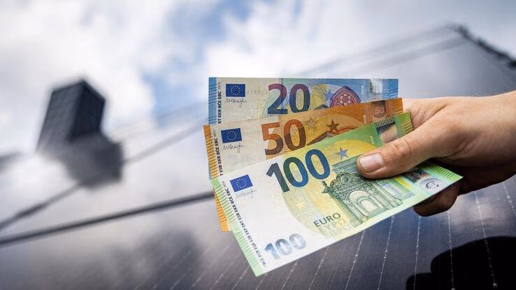 Das Foto zeigt drei Geldscheine vor einem Dach mit Solaranlage. Es ist ein 20 Euro, ein 50 Euro und ein 100 Euro Schein zusehen.