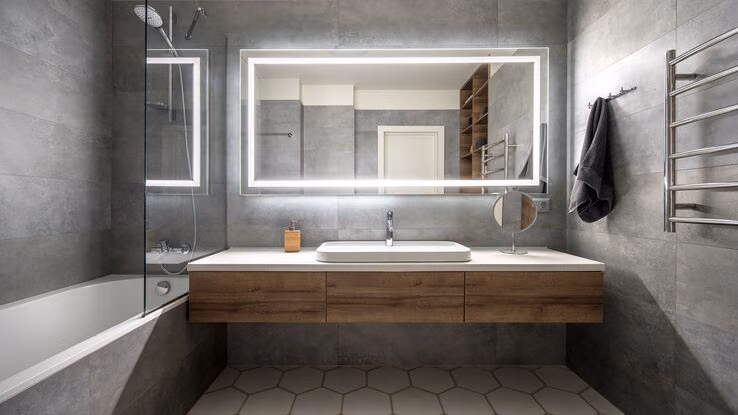 Ein Badezimmer mit genügend Ablagefläche und grauen Fliesen. Über dem Waschbecken hängt ein großer beleuchteter Spiegel. Auf der linken Seite ist eine Badewanne.