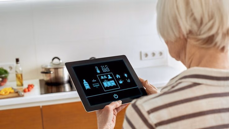 Eine ältere Frau hat ein Tablet in der Hand, mit dem sie ihr Zuhause steuern kann.