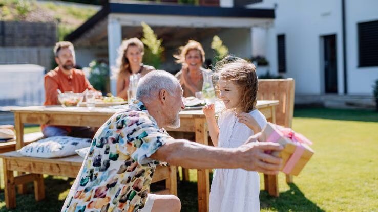 Das Foto zeigt eine Familie in einem Garten. Im Vordergrund übergibt der Großvater seiner Enkelin ein Geschenk. Im Hintergrund sitzen die Eltern des Kindes, sowie die Großmutter an einem Tisch.