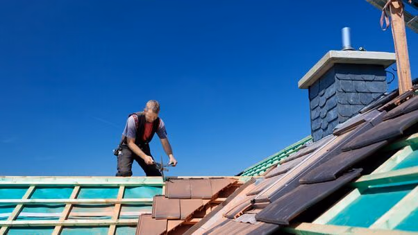 Ein Handwerker steht auf einem noch nicht fertig gedeckten Dach. Es sind überall noch die Streben zu sehen auf denen er nach und nach Ziegel anbringt.