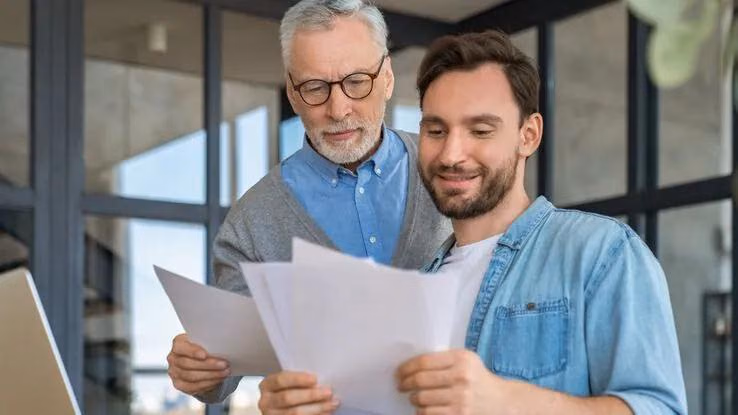 Das Foto zeigt einen äteren und einen jungen Mann. Möglicherweise Vater und Sohn. Sie halten Papiere in ihren Händen und lesen diese. Dabei wirken sie zufrieden was man an ihrem Lächeln erkennen kann.
