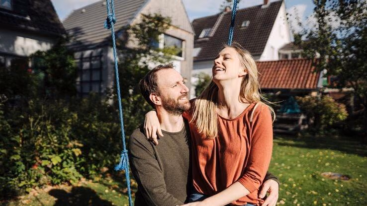 Das Bild zeigt ein Pärchen im Garten auf einer Schaukel. Die Frau sitzt auf dem Schoß des Mannes und beide lächeln sich an.