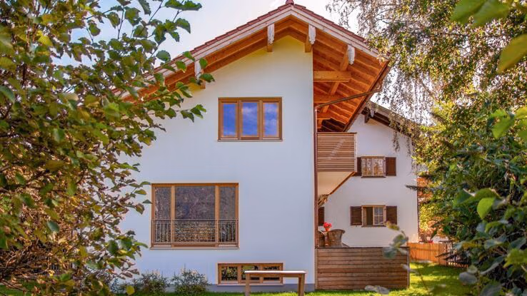 Das Foto zeigt ein großes Einfamilienhaus. Es wirkt als steht das Haus auf dem Land, da zusaätzlich noch ein großer Garten zu erkennen ist. Das Haus hat eine spezielle Form. Die eine Seite des Daches ist nämlich deutlich länger als die andere. Unter der rechten Hälfte des Dachs ist nämlich nur der Balkon und die Terasse. somit sieht das Haus aus, als sei es in der Mitte abgeschnitten worden. Zusätzlich ist im Hintergrund noch ein weiteres Haus zu erkennen.