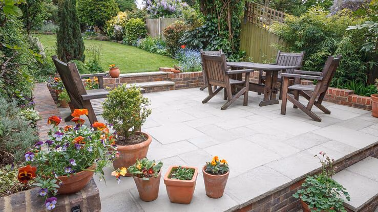 Das Foto zeigt einen Garten mit einer Steinterrasse. Auf der Terrasse stehen Holzmöbel, sowie einige Pflanzen und Blumen.
