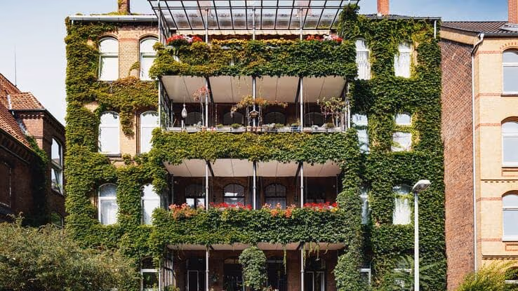 Das Foto zeigt ein Gebäude mit einer begrünten Fassade. Der Efeu überwächst das komplette Haus. Das Haus ist ein Altbau aus Ziegelsteinen. Zudem sind einige Balkone in der Mitte des Hauses zu sehen.