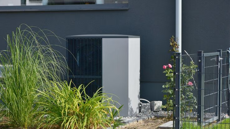 Abgebildet ist eine Wärmepumpe im Vorgarten eines Hauses. Sie steht direkt vor der Hauswand und ist von einem Strauch Gräser leicht verdeckt.