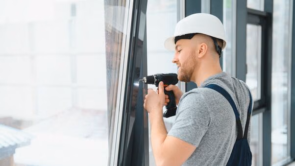 Ein Handwerker steht mit einem Akkuschrauber und einem Helm auf dem Kopf an einer Fensterfassade. Er scheint die Fenster auszutauschen.