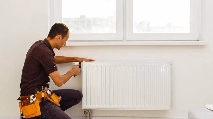 Ein Handwerker kniet an einer Heizung mit einem Werkzeuggürtel um die Hüfte und einem Werkzeug in der Hand. Er ist mit dem Heizungseinbau beschäftigt.