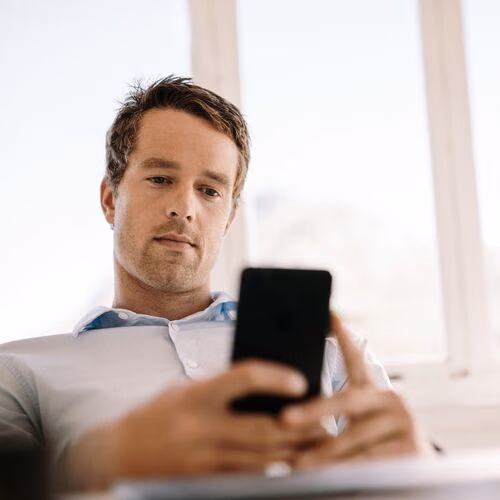 Person mit Smartphone in der Hand vor hellem Fenster im Innenraum.