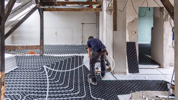 Das Bild zeigt einen Handwerker in einem Raum, der die Rohre einer Fußbodenheizung verlegt.