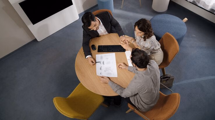 Das Foto zeigt ein junges Paar und einen Berater bei einem Gespräch. Die drei Personen sitzen an einem runden Tisch auf dem eine Maus, eine Tastatur und ein Blatt Papier liegt.