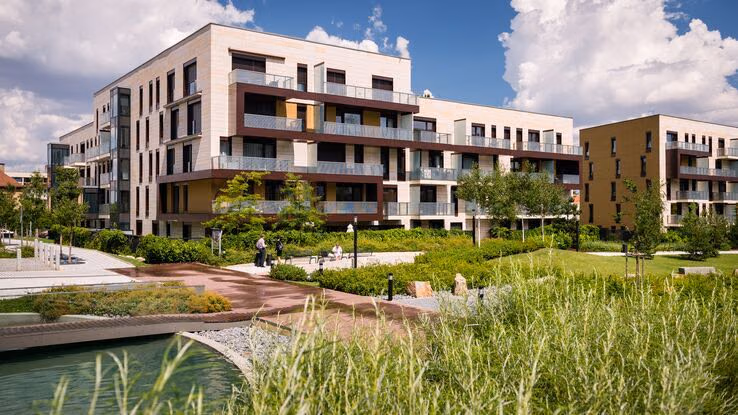 Das Foto zeigt eine moderne Wohnanlage. Es sind einige Wohnkomplexe, sowie ein kleiner Park mit einem Gewässer zu sehen.