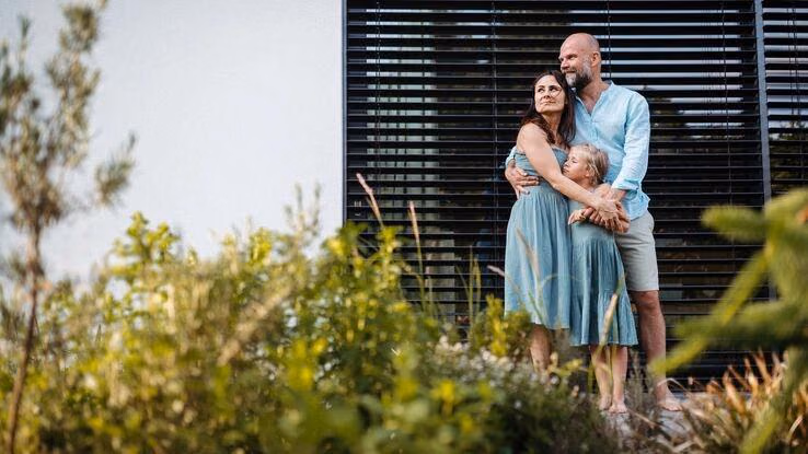 Eine kleine Familie steht Arm in Arm vor ihrer Immobilie.