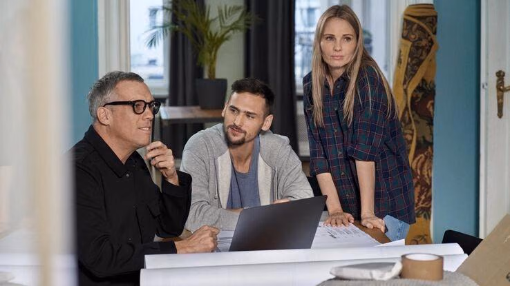 Drei Personen sind um einen Tisch versammelt. Ein Mann mit Brille hat einen Laptop aufgeklappt und der Mann rechts von ihm schaut ihn gespannt an. Die Frau steht neben ihnen und schaut in Richtung des Betrachters.