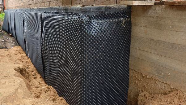 An einer Außenwand aus Holz sind Plastikpaneelen mit Noppen angebracht. Der Boden ist mit einer Schicht Sand bedeckt.