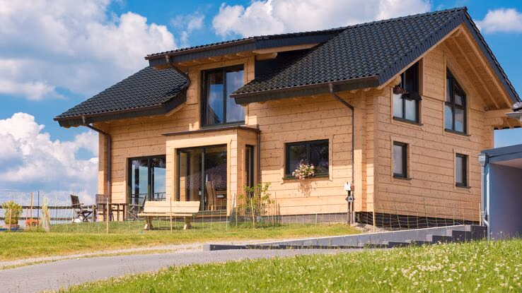 Das Foto zeigt ein Einfamilienhaus aus Holz. Es hat eine Dachgaube in der Mitte des Daches und große schwarze Fenster. Das Haus wirkt sehr neu, was man auch am provisorischen Zaun erkennen kann.