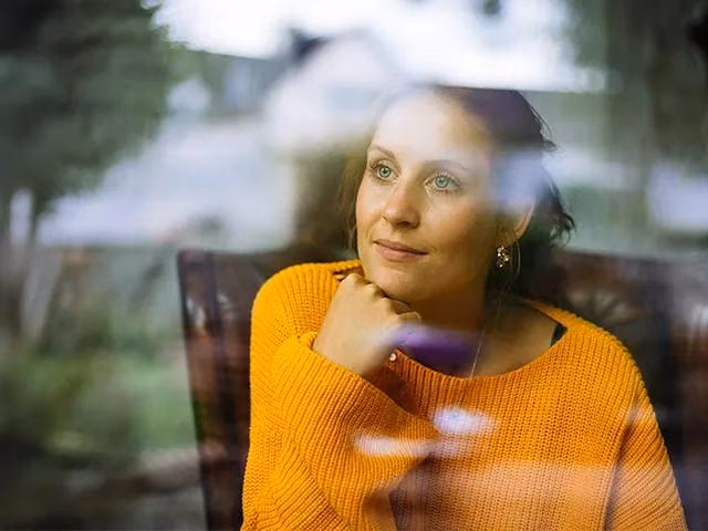 Person sitzt in einem Wohnraum am Fenster und blickt nach draußen.