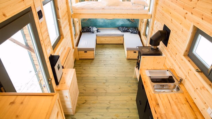 Das Foto zeigt ein Tiny House beziehungsweise Caravan von innen. Auf der rechten Seite ist eine Küchenzeile zu erkennen. Außerdem hat das Tiny House noch einen weiteren Raum im Erdgeschoss. Zudem lässt eine Holzleiter vermuten, dass im ersten Stock ein weiterer Raum ist.