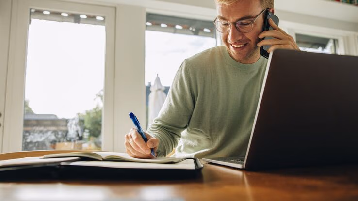 Ein Mann telefoniert und schreibt etwas auf, während er am Laptop Immobilienrecherche betreibt.