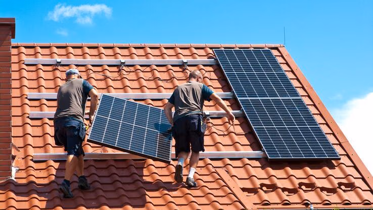 Zwei Handwerker heben gemeinsam ein Solarpanel auf das Dach eines Gebäudes, um es dort zu montieren.