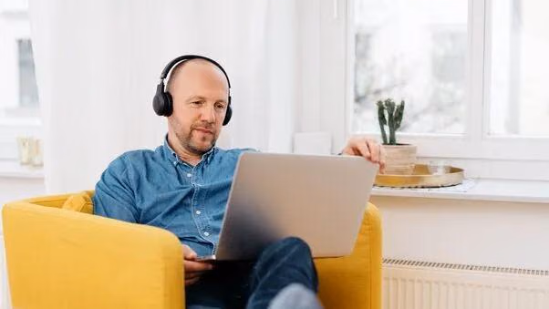 Ein Mann sitzt in einem Sessel, hat einen Laptop in der Hand und Kopfhörer auf.