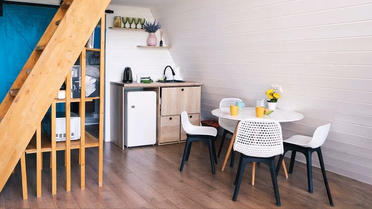 Das Foto zeigt ein Zimmer mit einer kleinen Küchenzeile und einem Esstisch. Außerdem eine steile Holztreppe, die in ein höheres Stockwerk führt.