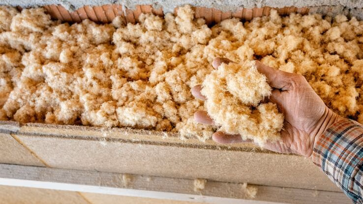 Eine Hand greift in die Holzfaserdämmung, welche zwischen Ziegeln und einer Holzplatte gefüllt ist.
