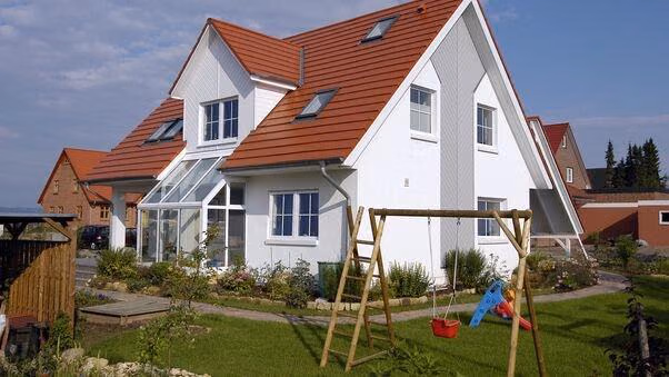 Das Foto zeigt ein Einfamilienhaus mit Garten und Schaukel. Im Hintergrund sind noch zwei weitere Einfamilienhäuser zu sehen. Die Häuser stehen wahrscheinlich auf dem Land.