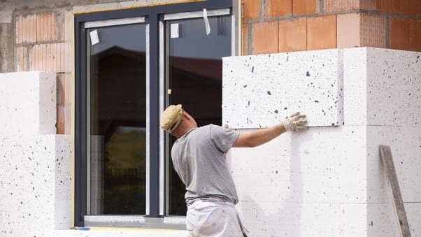 Ein Handwerker mit einem kurzen T-Shirt und Handschuhen befestigt eine Dämmstoffplatte an einer Hauswand.