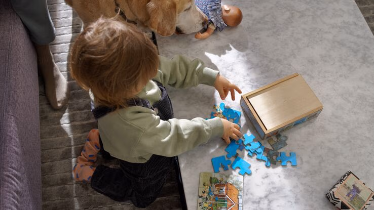 Man sieht ein Kind von oben beim Spielen mit Puzzleteilen. Neben dem Kind steht ein Hund.