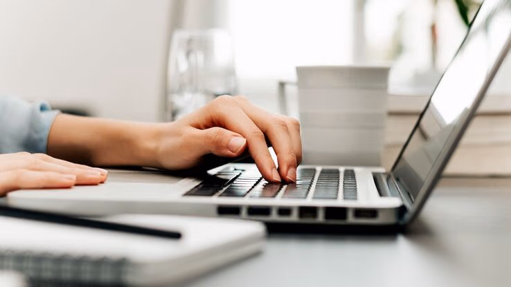 Das Foto zeigt eine Nahaufnahme einer Hand die auf der Tastatur eines Laptops liegt. Der Laptop steht auf einem Schreibtisch. Außerdem liegt ein Notizbuch und ein Stift im Vordergrund.
