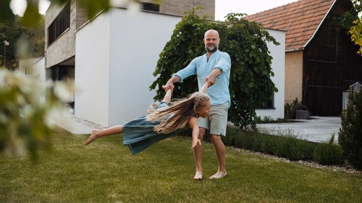 Ein Vater hat seine Tochter an einem Hand und einem Fuß und schwingt sie durch den eigenen Garten.