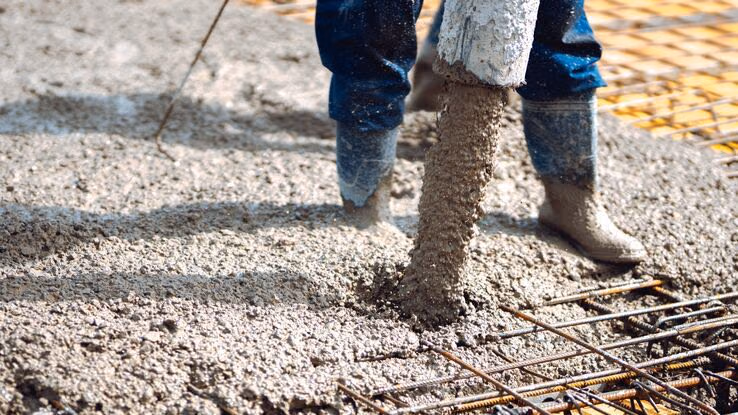 Das Foto zeigt einen Bauarbeiter beim Betonieren einer Bodenplatte. Er steht mit Gummistiefeln im Beton und gießt den Zement mit Hilfe eines Schlauchs auf das Eisengitter.