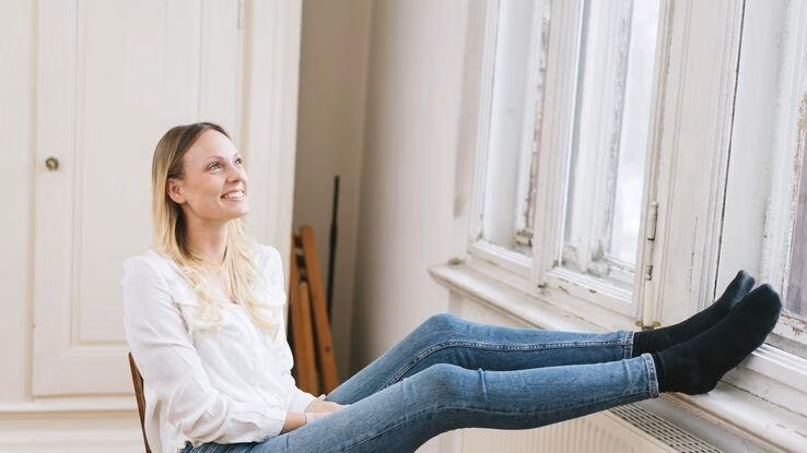 Eine Frau schaut in ihrer Altbauwohnung aus dem Fenster und hat die Füße auf der Fensterbank abgelegt.