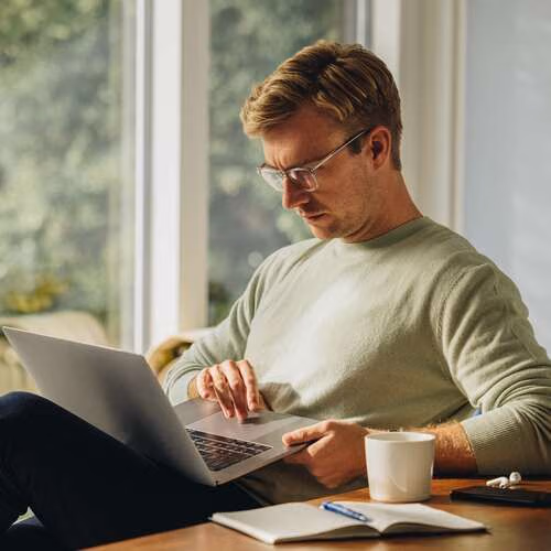 Ein Mann sitzt mit einer Tasse Kaffee am Laptop und nutzt Interhyp Home.