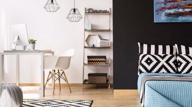 Zimmer mit Schreibtisch und Bett. Außerdem ist ein offenes Regal mit Deko darin zu sehen.