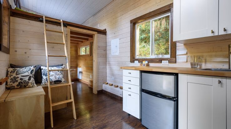 Das Foto zeigt ein Tiny House von innen. Es beinhaltet eine kleine Küchenzeile, eine Sitzbank, sowie ein weiteres Zimmer im Hintergrund.