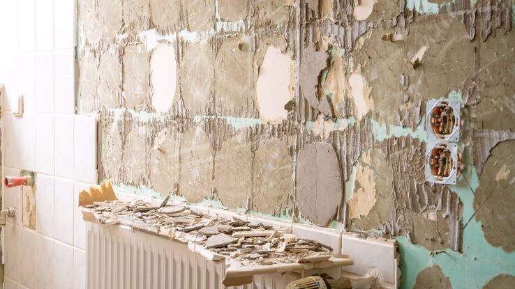 Es ist eine Wand abgebildet, von der die Fliesen abgemacht wurden. Auf der Heizung liegen die Überreste. Hinten ist noch ein Teil der Fliesen an der Wand zu sehen.