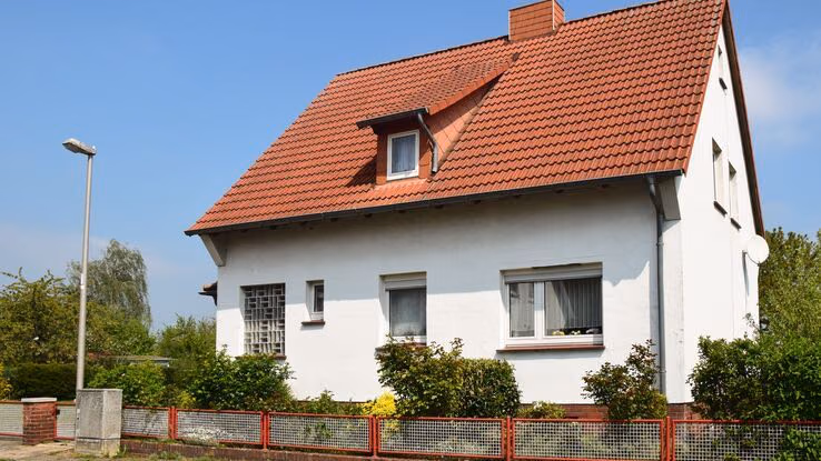 Das Foto zeigt ein älteres Einfamilienhaus. Die Wände des Hauses sind weiß und die Ziegel sind klassisch rot. Außerdem hat das Haus eine Dachgaube auf der Seite. Das Haus hat einen Garten mit grünen Pflanzen. Zwischen Haus und Straße ist ein weiß - roter Zaun.
