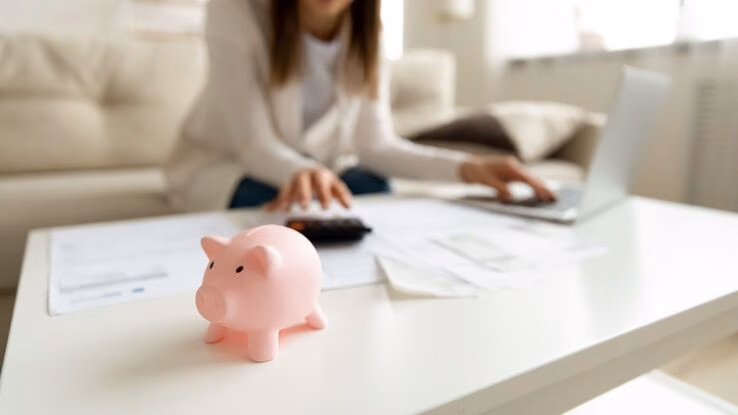 Eine Frau arbeitet an ihrem Laptop auf dem Sofa. Im Vordergrund steht ein kleines Sparschwein.