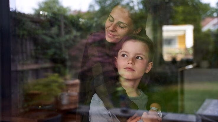 Eine Mutter umarmt ihren kleinen Sohn von hinten, während er aus dem Fenster schaut.