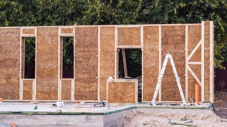 Das Bild zeigt eine Baustelle mit einer teilweise errichteten Wand aus Holzrahmen und Strohballen.