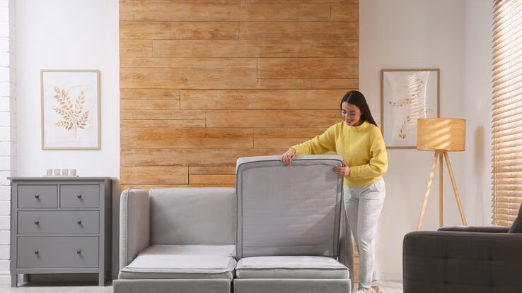 Das Foto zeigt eine Frau die eine Couch als Bett umbaut. Außerdem sind in dem Raum ein Regal, eine Stehlampe, sowie ein Sessel zu sehen.