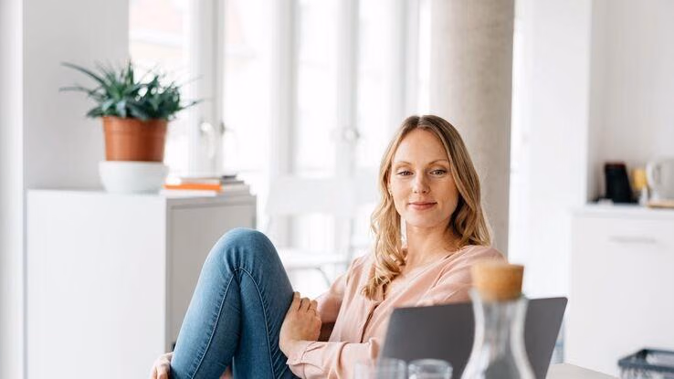 Das Bild zeigt eine Frau, die in einem hellen, modernen Raum sitzt, die Beine überkreuzt hat und einen Laptop auf dem Tisch vor sich stehen hat. Im Hintergrund steht eine Topfpflanze auf einem weißen Schrank, zusammen mit einigen Büchern.
