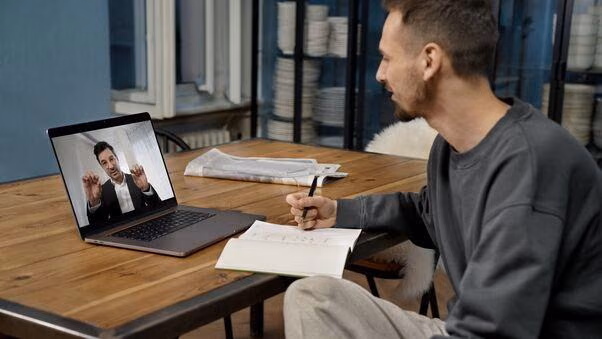Ein Mann sitzt an einem Schreibtisch und wird Online Beraten. Auf dem Laptop erkennt man einen Berater im Anzug. Der Mann macht sich in auf einem Schreibblock notizen.
