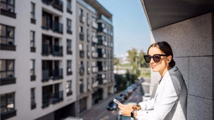 Eine Frau steht auf dem Balkon von einem Bürogebäude und hat ein Smartphone in der Hand.