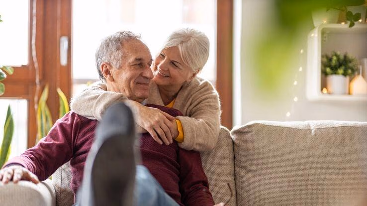 Das Foto zeigt ein älteres Pärchen. Der Mann sitzt auf einer Couch und die Frau umarmt ihn von hinten.